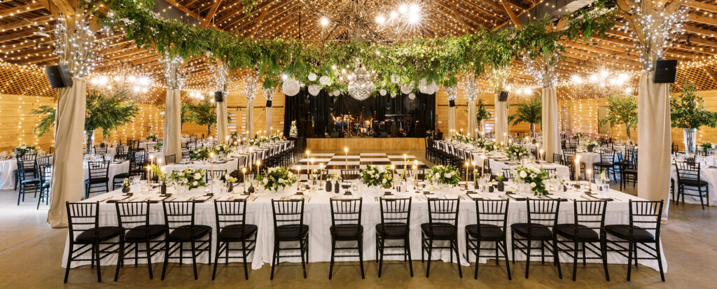 A decorated barn venue set for a wedding, featuring long banquet tables with white linens, black chairs, greenery, candles, and string lights hanging from a wooden ceiling. A stage with musical instruments is in the background.