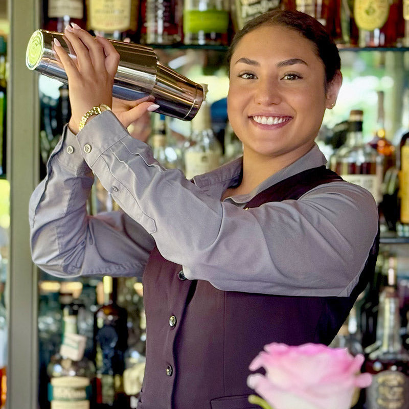 A smiling bartender wearing a vest and gray shirt shakes a cocktail shaker behind a bar stocked with various bottles, with a pink flower in the foreground.