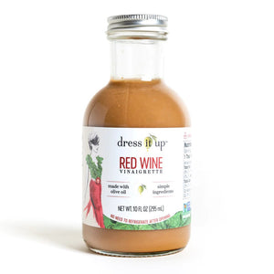 A bottle of "DRESS IT UP DRESSING - RED WINE" from the brand "DRESS IT UP DRESSING," featuring an illustrated woman in a green top and red skirt on the label. Made with olive oil and simple ingredients, it has a net weight of 10.1 fl oz (295 ml).