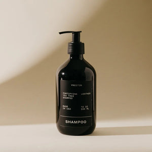 A black bottle of PRESTON - SHAMPOO with a pump dispenser stands against a beige background. The label emphasizes features like "Amber," "Made in USA," and "16 fl oz / 500 ml." Shadows are cast to the left, enhancing the minimalist aesthetic. This sulfate-free shampoo is perfect for volumizing and moisturizing.