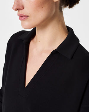 A person with dark hair styled in a neat updo is wearing the SPANX - AirEssentials Split Neck Polo Top in black, paired with matching black pants. They rest one hand on the back of their neck and gaze to the side against a plain white background, exuding an air of luxurious comfort.