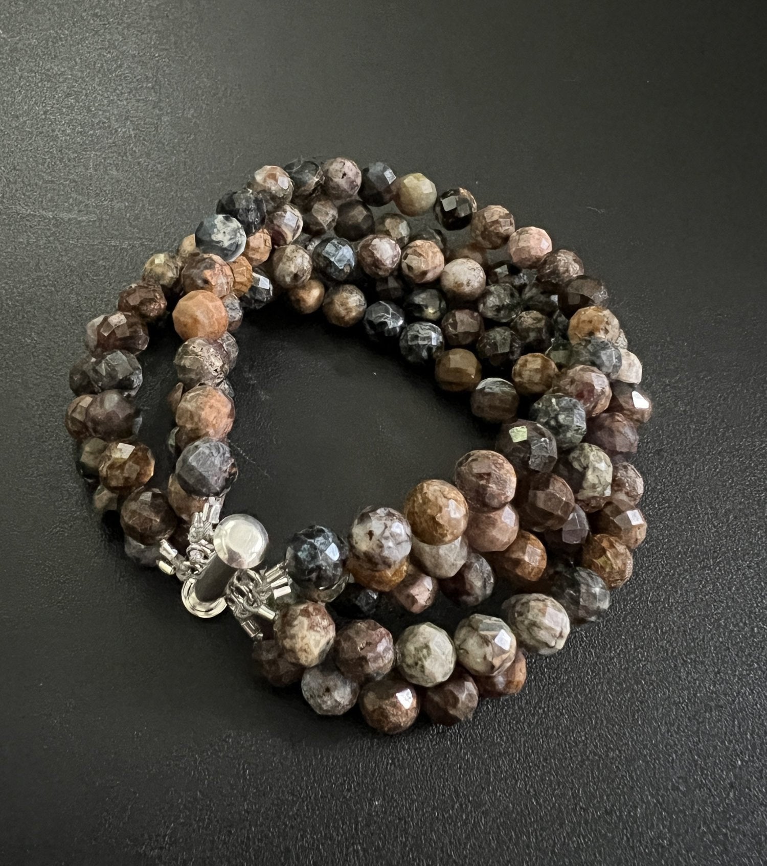 The ANN LIGHTFOOT - FIVE STRAND 6MM FACETED PIETERSITE BRACELET, crafted by Ann Lightfoot, is showcased against a black background. The bracelet features faceted pietersite beads in an array of brown and gray shades, displaying a marbled look, and is secured with a stainless steel clasp.