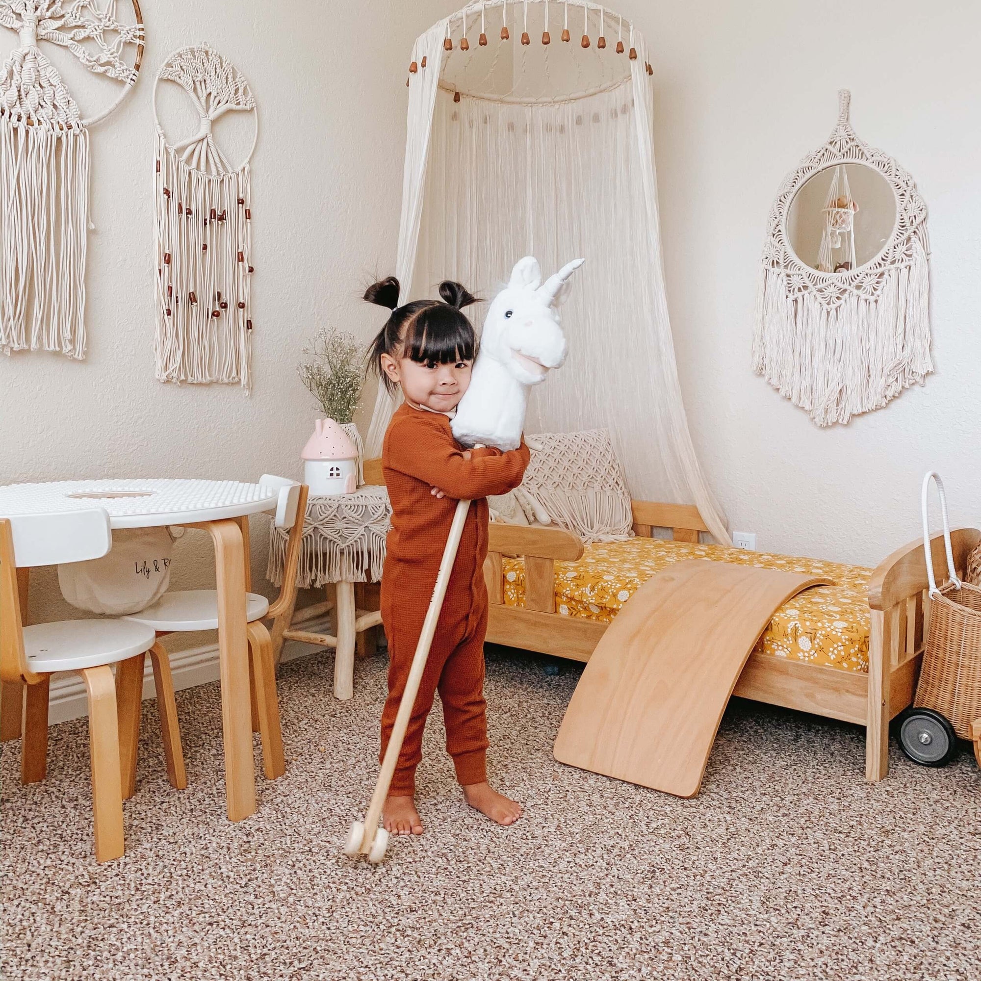 A young child in a cozy, bohemian-style bedroom holds the LITTLE UNICORN RIDER by LILY & RIVER, sparking their imagination. The room showcases children's products such as a small wooden bed with a canopy, a table and chairs, and wall decorations made of macrame. Dressed in brown with hair styled in two buns, the child enjoys playful physical activity.