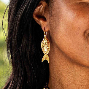 Close-up of a person wearing a pair of CAPUCINE DE WULF - POISSON DROP EARRINGS. The person's fingers, with light-colored nails, are touching their long dark hair. The image highlights the intricate design of the 18K Gold IP earring by CAPUCINE DE WULF and the person's ear and neck area.