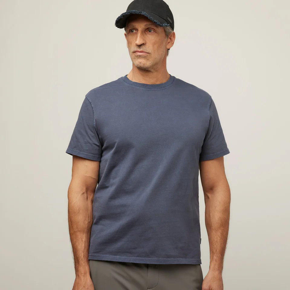 A man in a TILLEY MID WEIGHT SUNWASHED T-SHIRT IN MARINE NAVY, gray pants, and a black cap stands against a plain light background, gazing slightly to the side with a neutral expression.