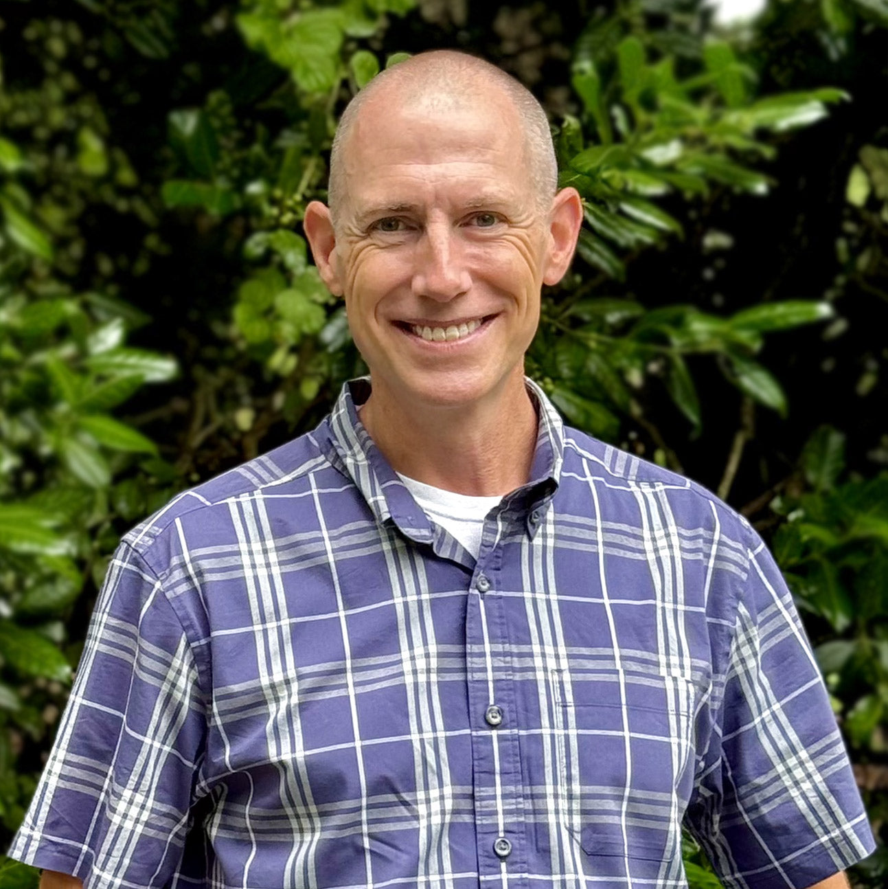 A smiling, bald man wearing a blue and white plaid shirt stands outdoors in front of lush green foliage.