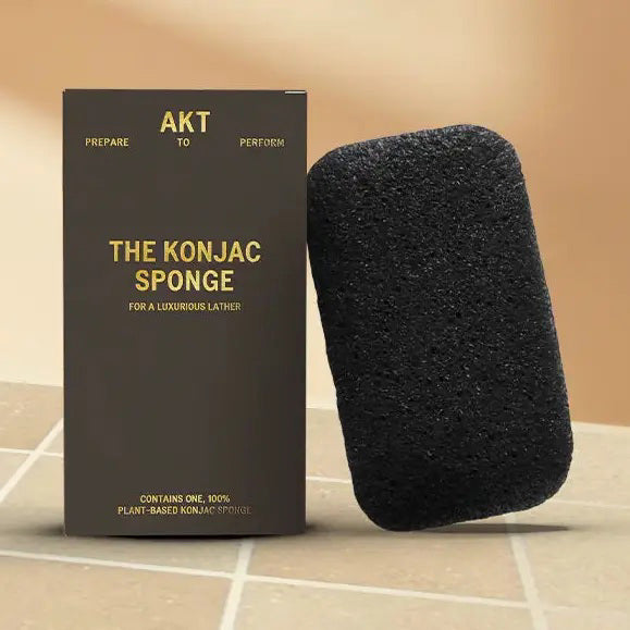 A black rectangular AKT LONDON - KONJAC FACE AND BODY SPONGE with activated charcoal leans against a dark brown AKT LONDON box featuring gold lettering, set on beige tiles with a neutral backdrop.