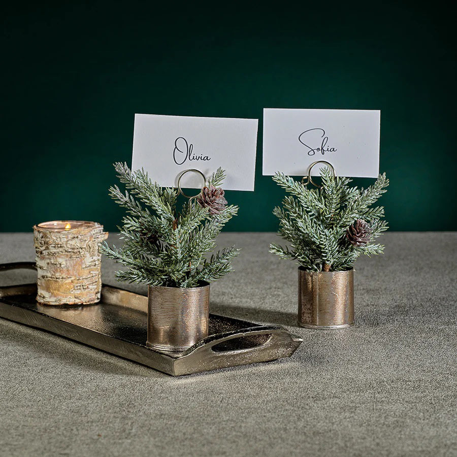 Two ZODAX PINE IN SILVER BUCKET PLACE CARD HOLDERS, featuring place cards with the names "Olivia" and "Sophia," adorn a gray surface. Adjacent to them, a lit candle in a 3 in x 5.75 in birch-patterned holder sits on a metal tray against a dark green backdrop.