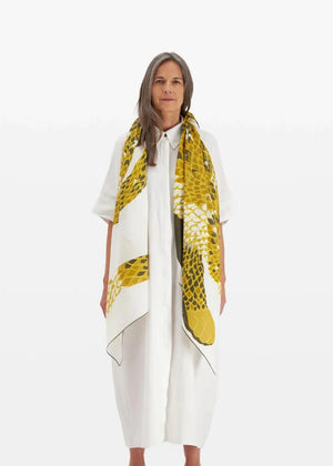 A woman with long, grey hair is wearing a white, loose-fitting dress and has an INOUI EDITIONS - NEOFELIS cotton and silk scarf from INOUI, featuring a snake pattern in yellow, draped around her shoulders. She stands against a plain, white background, looking directly at the camera.