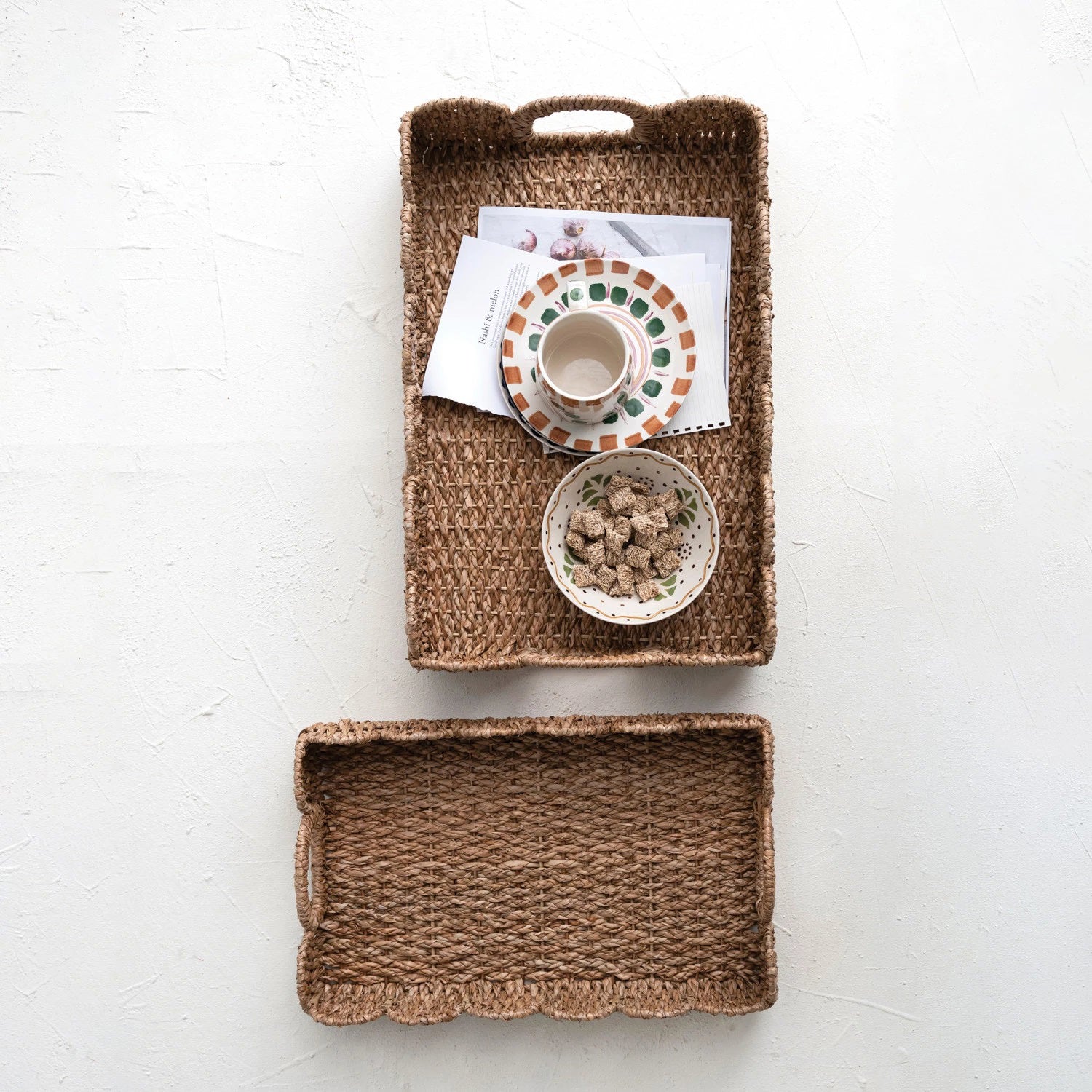 Two woven rectangular baskets with scalloped edges and sturdy handles sit side by side. The baskets feature a natural, rustic look with a textured finish. The left basket, slightly smaller than the right, adds charm reminiscent of the LARGE BRAIDED BANKUAN TRAY WITH HANDLES by CREATIVE COOP.