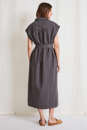 A woman is showcasing the APIECE APART - CAMI MAXI DRESS, a sleeveless, button-down creation in dark gray, made from organic cotton. It features a matching tie belt for added style. With its relaxed fit and collar design, she stands against a plain background, displaying her right side.