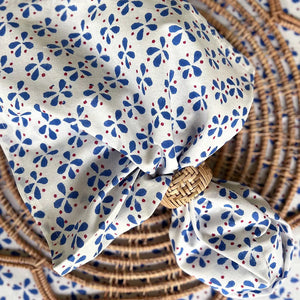 The CHRISTINA DICKSON HOME - PETALS COTTON NAPKIN showcases a blue and red floral pattern on white fabric, crafted in India. Made from 100% cotton, it is knotted over a rattan circle, creating an elegant and textured drape.