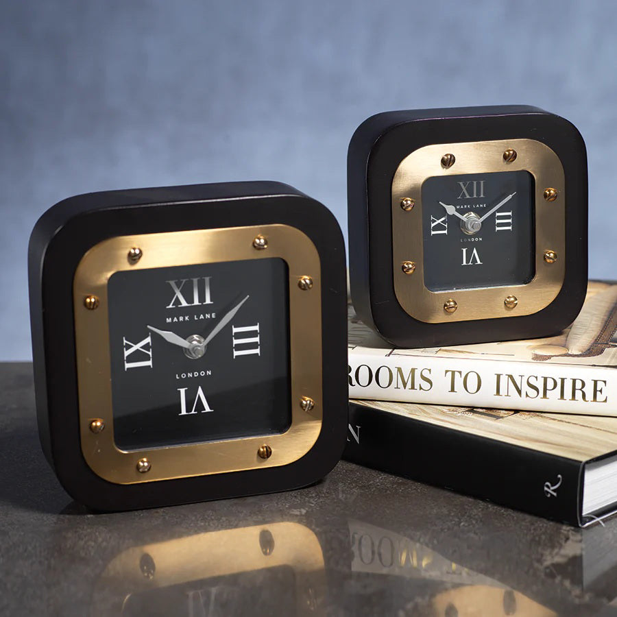 Two MARK LANE TABLE CLOCK - LARGE by ZODAX, square in shape with black and gold iron finish and Roman numerals, are displayed—one on a table, the other atop &quot;Rooms to Inspire&quot; books. The clocks show different times.