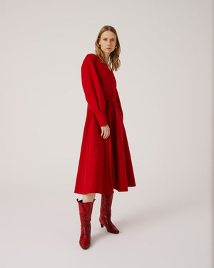 A woman with long, light brown hair stands against a plain background. She is wearing the SFIZIO long sleeve v-neck flare skirt dress, featuring a tie waist. Looking to her left with a calm expression, she has hoop earrings and exudes minimalist style.