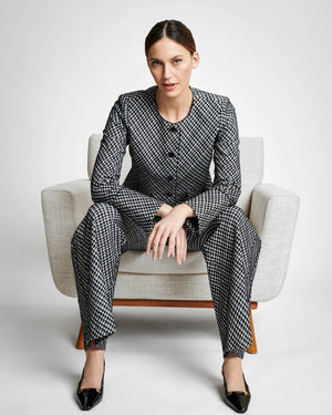 A person with long hair sits casually on a light-colored chair wearing the SPANX PONTE JACQUARD JACKET, which is a black and white checkered suit adorned with gold-finished buttons. They are gazing to the side, with one arm resting on the chair's armrest and the other in their lap.