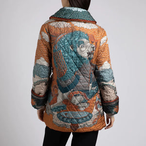 A person wears the SABINA SAVAGE - THE SNOW LION QUILTED JACKET from SABINA SAVAGE ENGLAND, featuring a colorful, intricately designed nature-themed pattern with trees, sky, and animals. Reminiscent of a reversible silk twill jacket, it features a wide collar, contrasting orange inner lining, and large pockets. The person is shown from the shoulders down.