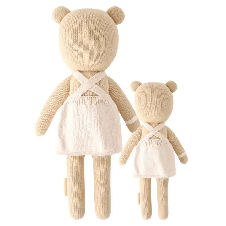 Two beige, hand-knit Goldie the Honey Bears from CUDDLE & KIND, one larger at 20 inches and one smaller, are standing side by side. Both bears are wearing white aprons that cross at the back. Made from safe, non-toxic materials, these fair trade dolls have simple features with no visible faces or distinguishing characteristics.