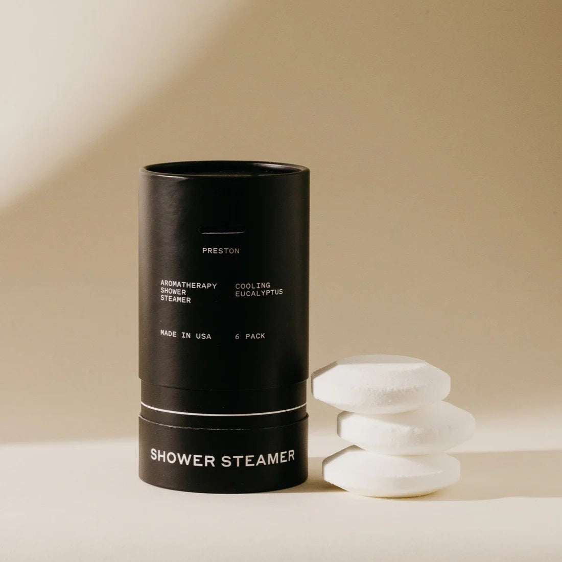 A cylindrical black container labeled &quot;PRESTON - SHOWER STEAMER COOLING EUCALYPTUS&quot; and &quot;Cooling Eucalyptus&quot; with an open lid stands upright. Three white aromatherapy tablets rest beside it on a neutral background. The container also reads &quot;Made in USA&quot; and &quot;6 Pack.