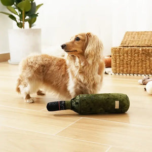 A curly-haired dog wearing a light blue bandana relaxes on a yellow plaid blanket outdoors. Surrounding the dog are various toys, including a plush cheese and LAMBWOLF COLLECTIVE's MERLOT DOG TOY. In the background, trees and a building can be seen.