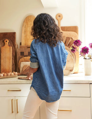 A person with curly hair stands in a kitchen, leaning back against a counter with hands resting on the surface. They wear an Eileen Relaxed Button Up Vintage Indigo Denim shirt from FRANK & EILEEN with rolled-up sleeves and white pants. The counter has a collection of wooden cutting boards and a vase with purple flowers.