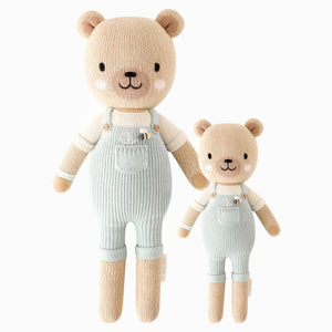 Two CUDDLE & KIND Charlie the Honey Bears are shown standing side by side. Both handmade bears are wearing light blue overalls and have beige fur with cute embroidered facial features. The larger 20" bear is on the left, and the smaller bear on the right. These adorable hand-knit dolls make perfect baby shower gifts, set against a plain white background.