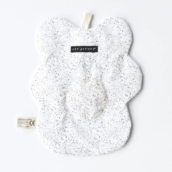 The WEE GALLERY - EGG CRINKLE TOY by WEE GALLERY is a white baby crinkle toy in the shape of a flower with a yellow circle at the center that features a smiling face with closed eyes. Made from organic cotton, it comes with a brown tag inscribed &quot;Baby Crinkle Toy - Organic Crinkle Toy.&quot; The toy is displayed hanging on a white hook.