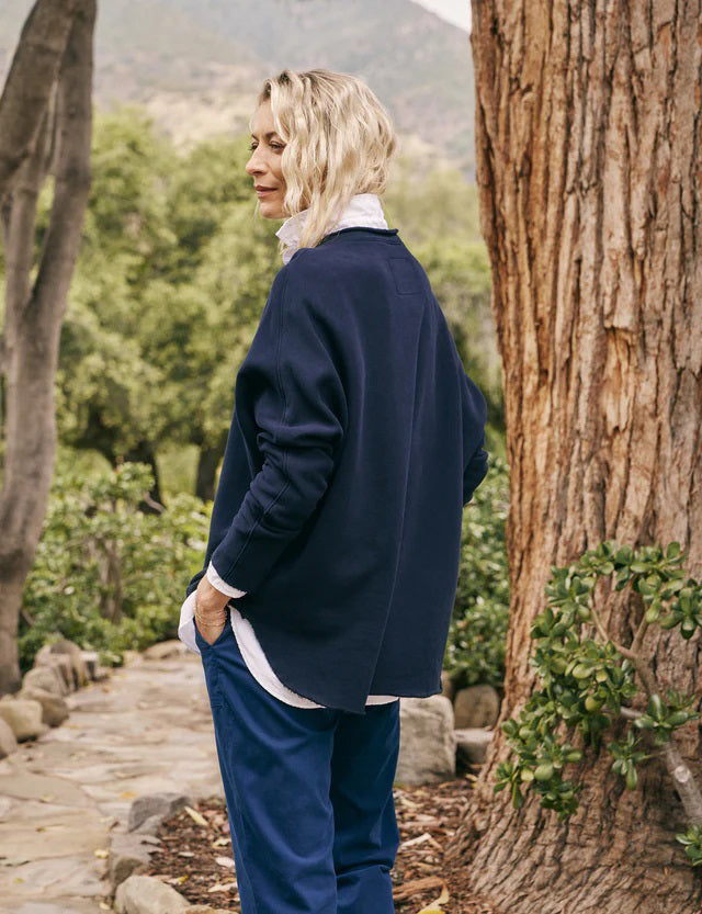 A person with long, wavy blonde hair is standing outdoors near a large tree, wearing the FRANK & EILEEN ANNA LONG-SLEEVE CAPELET in navy over a white shirt and blue pants. The background showcases green foliage and a stone path. The person is smiling and gazing to the side.