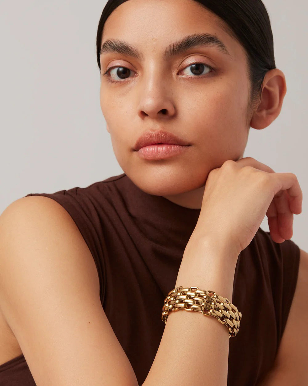 A person with long dark hair pulled back is wearing a brown sleeveless top and a JENNY BIRD - PENELOPE MEDIUM GOLD BRACELET by JENNYBIRD, featuring a foldover clasp. They rest their chin on their hand with a neutral expression against a plain background.