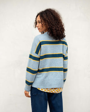 A person with curly hair stands against a light wall, wearing the ultra-soft MERSEA - KEEPER CREWNECK SWEATER in light pink with black and red stripes, paired with blue jeans. The individual faces away, looking to the left.