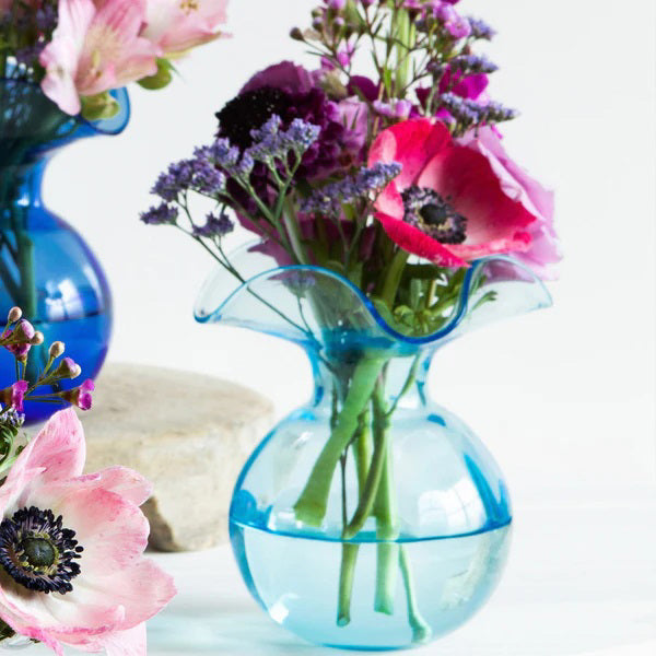 The VIETRI Hibiscus Bud Vase in Aqua features a sculptural design with a curvy, wide top and rounded body. Its translucent blue material reflects light beautifully, evoking the elegance of a blooming hibiscus flower and showcasing the artistry of Tuscan artisans.