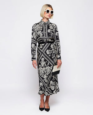 A woman with blonde, chin-length hair is wearing the MIRTO 1956 - BLACK AND WHITE PRINT FLUID BLOUSE. Embracing the intricate flower squares patchwork print of MIRTO fashion, she stands neutrally against a plain white background.