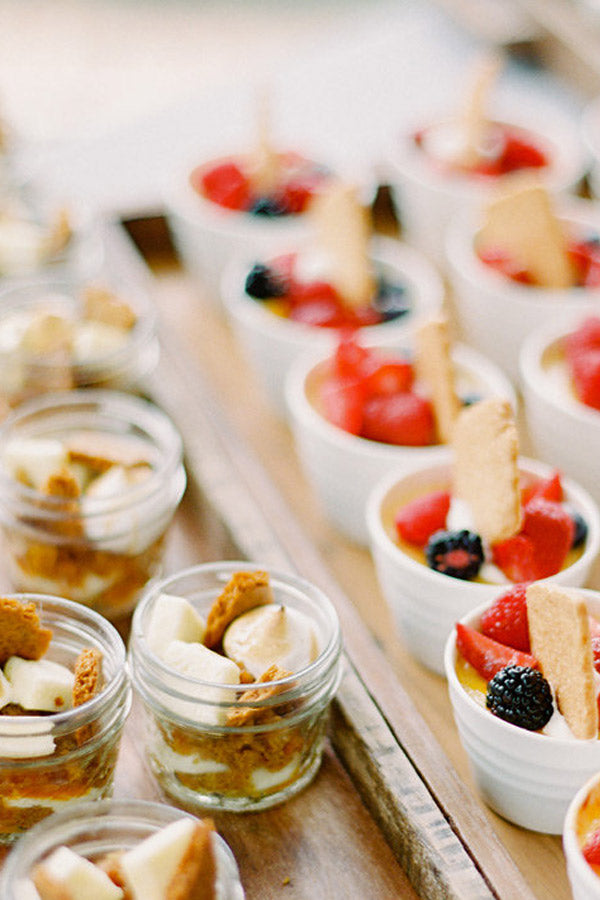 Individual dessert cups filled with layered ingredients, including cookies or biscuits, fruit such as strawberries and blackberries, and creamy fillings, arranged neatly on a wooden tray.