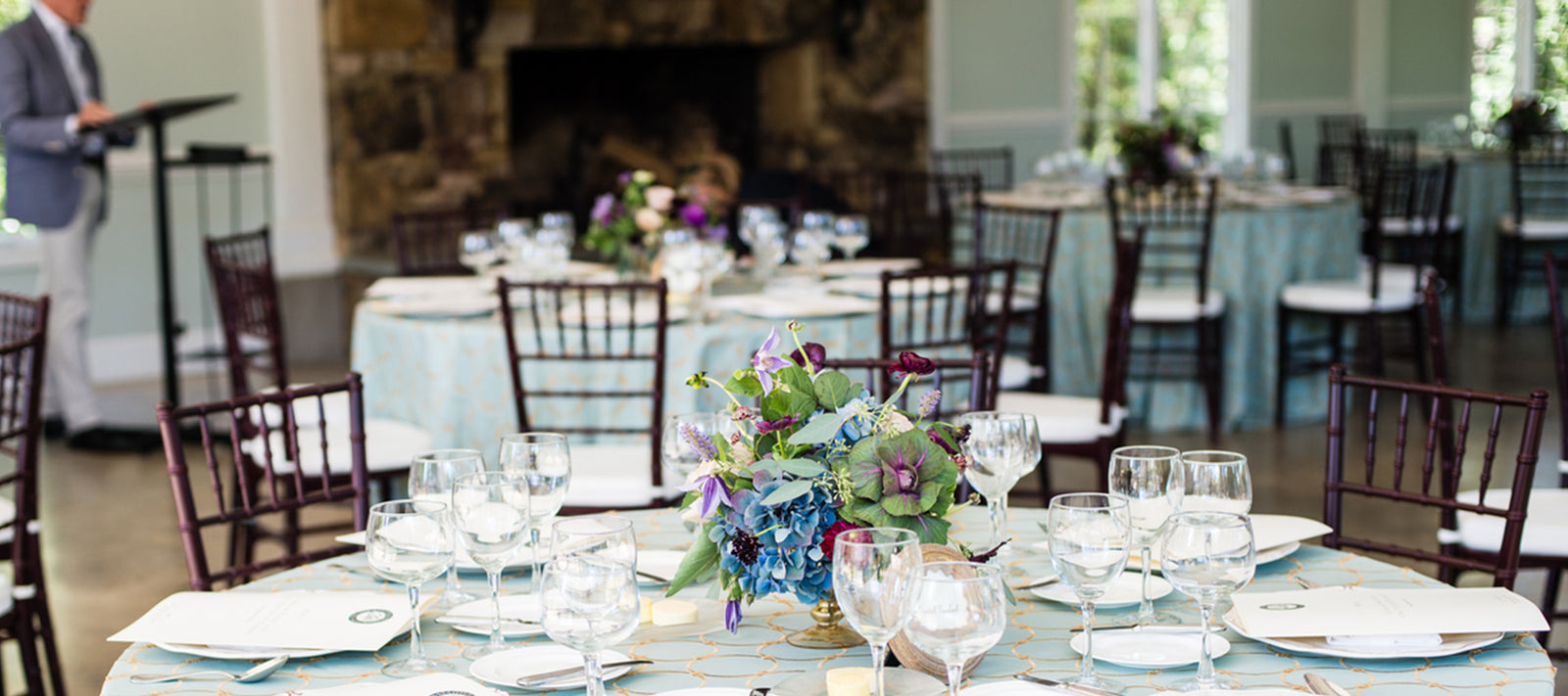 Elegant dining tables with floral centerpieces, neatly arranged glassware, and menu cards are set in a bright, airy room. A person stands at a podium near a stone fireplace in the background.