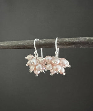 The ANN LIGHTFOOT Pale Pink Freshwater Pearl Cluster Earrings elegantly hang on a dark rod, featuring clusters of small pearls with a smooth, shiny surface in a floral design attached to sterling silver earwires, adding sophistication.