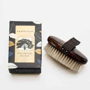 A rectangular box labeled "BAUDELAIRE - BEECHWOOD 5\" BODY BRUSH" with a tree illustration stands upright on a white surface. Next to it lies a beechwood body brush featuring natural horsehair bristles and a black hand strap. Both items evoke the essence of traditional Japanese rituals against the plain white background.