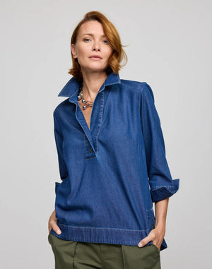 A person with short hair sits on a stool, facing away from the camera. They wear the breathable HINSON WU AILEEN 3/4 sleeve top in denim, featuring buttons down the back, paired with green pants.