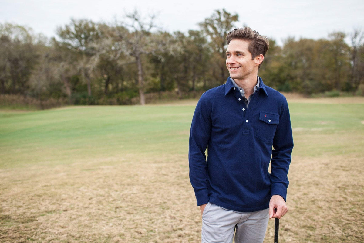 A man wears the CRIQUET Long Sleeve Players Polo Shirt in Peacoat, made from soft Peruvian Pima cotton with a buttoned collar and chest pocket. He stands with one hand in his jeans pocket against a white background.
