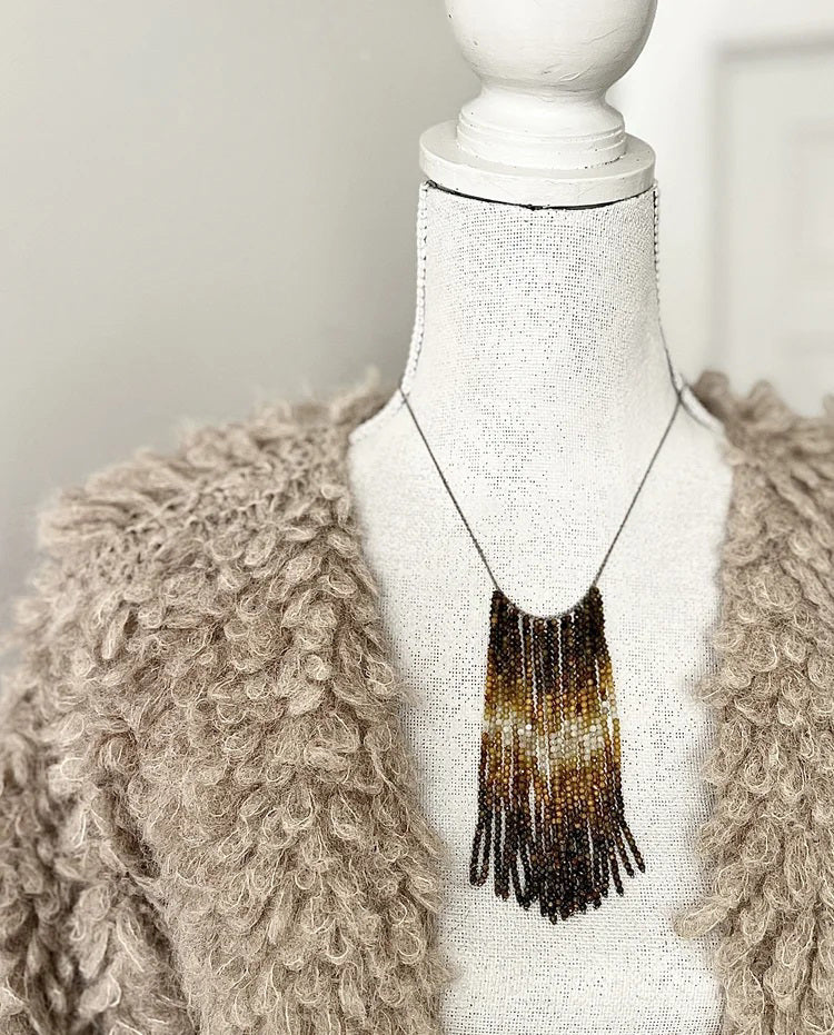 A mannequin wears a textured, curly taupe jacket and the ANN LIGHTFOOT Tiger Eye and Quartz Ombre Cascade Necklace, featuring dramatic dark beaded fringes in a triangular shape. The softly blurred background is neutral.