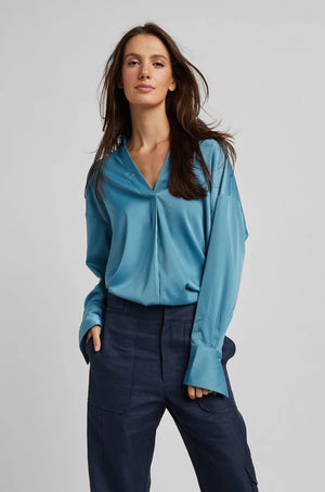 A person with long brown hair is standing with their back partially turned, wearing the ADROITE ATELIER - KAY POPOVER SATIN BLOUSE, an oversized white V-neck satin blouse from ADROIT ATELIER, paired with light blue jeans. The person is also wearing large, ornate earrings. The background is plain and light-colored.