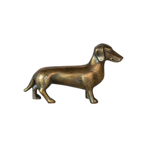 A polished CAST ALUMINUM DACHSUND by CREATIVE COOP stands atop an open book, making the perfect dachshund accent for dog lovers decor. The book lies on a light-colored surface, with text and illustrations visible on the pages. The background is blurred to focus attention on the vintage brass-like finish of the figurine and the book.