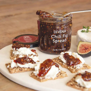 A jar of DIVINA - CHILI & FIG SPREAD from GOURMET FOOD SOLUTIONS features a black lid and a transparent body, showcasing the dark, chunky, sweet and spicy spread inside. The label on the front reads “DIVINA - CHILI & FIG SPREAD” and indicates a net weight of 9 oz (255 g). Ideal for pairing with cheese, it is certified Non-GMO Project Verified.