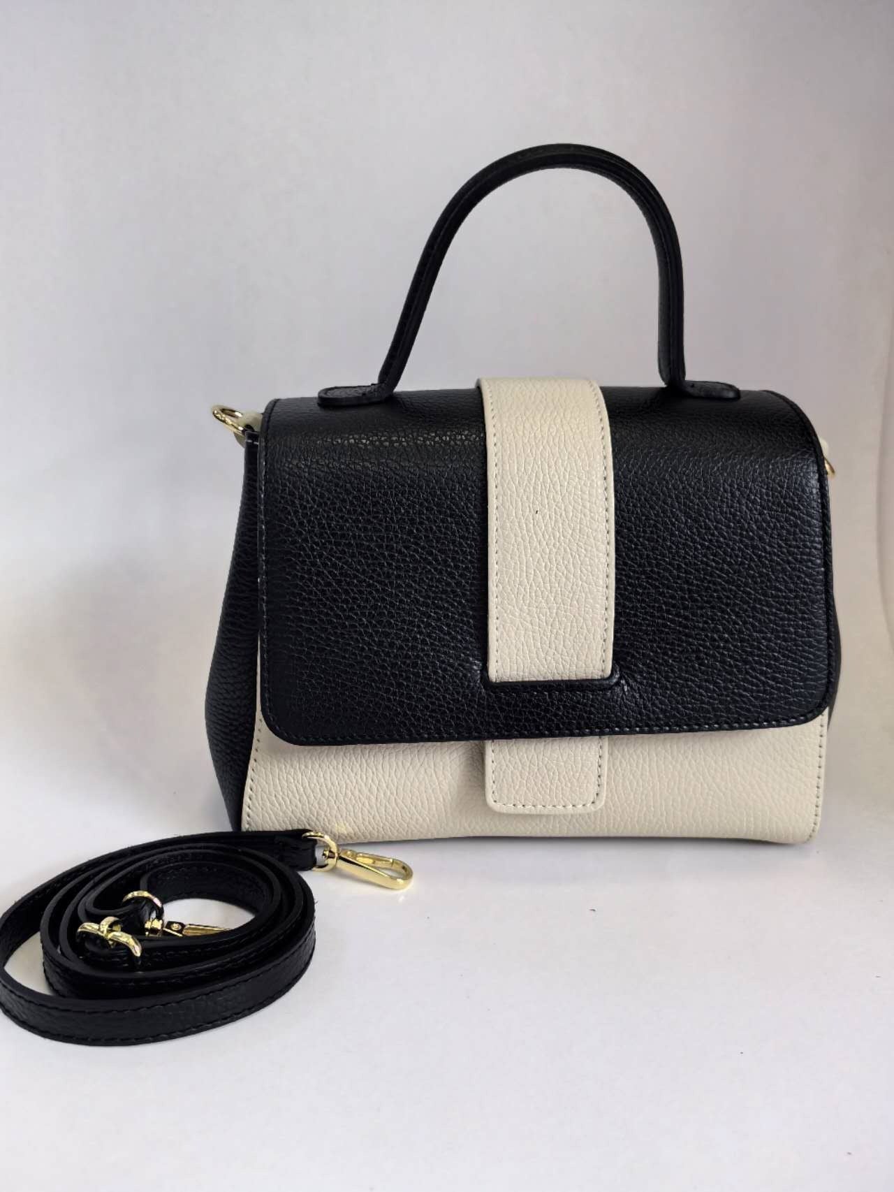 The TWO TONE LEATHER HANDBAG by MONDO BORSE is a black and white leather accessory featuring a top handle and a fold-over flap. The Italian-made handbag is predominantly black with a distinctive white stripe on the front flap. Accompanying it is a detachable, adjustable black shoulder strap adorned with gold hardware, all set against a plain white background.