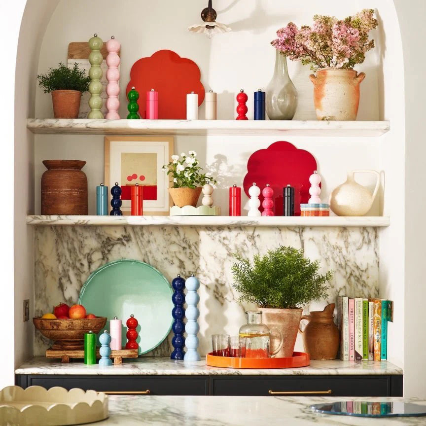 A decorative kitchen shelf with various colorful items including candles, potted plants, a bowl of fruit, books, a green plate, a red tray with a pitcher and glasses, and a vase of flowers. The ADDISON ROSS LONDON - GRANDE SALT OR PEPPER MILL 35CM by ADDISON ROSS adds to the charm against the white marble-patterned backdrop.