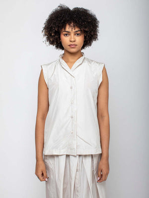 A curly-haired woman models the KATHARINA HOVMAN - SLEEVELESS TOP, standing against a plain white background. She wears a V-neck, white button-up blouse and gazes directly at the camera with a neutral expression.