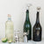 Three bottles featuring animal-shaped stoppers, including the CAST ALUMINIUM DOG BOTTLE STOPPER by CREATIVE COOP, rest on a marble surface with a sliced lime. They are crafted from green and clear glass, showcasing shiny metal and cast aluminum finishes.