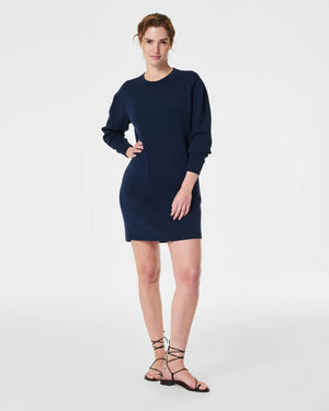 A woman is wearing the SPANX AirEssentials Crew Neck Dress, a black, long-sleeve, knee-length dress from the SPANX brand, with her hands in the functional pockets. She is looking to the side and has long, straight hair. Paired with white sneakers, she stands against a plain white background in this light-as-air fabric attire.