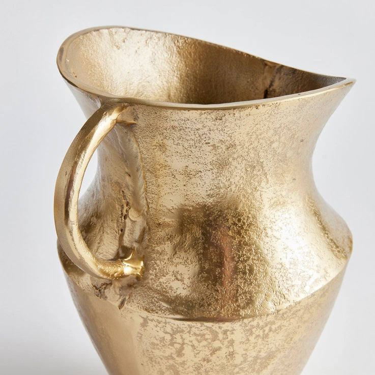 The ELAINA DECO PITCHER by NAPA HOME AND GARDEN features a golden, textured cast aluminum design with a large, curved handle and a slightly flared rim. Its surface boasts a matte, hammered finish that offers a rustic yet elegant look, ideal for adding charm to any transitional space. The decorative pitcher is showcased against a plain white background.