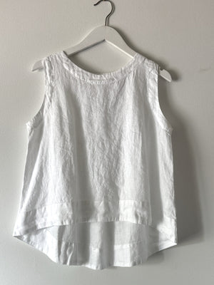 The CUT LOOSE - HI LOW TANK, a sleeveless white linen blouse with a round neckline, is displayed on a white hanger against a light gray wall. Perfect for summer wear, this breathable top by CUT LOOSE features a loose fit and an elegant high-low hem that is shorter in the front and longer in the back.