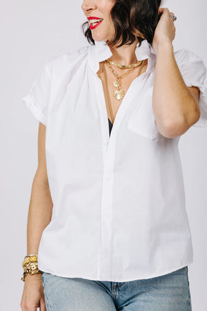 A person wearing the KMJ - QUARANTINI SHORT SLEEVE SHIRT with the bottom two buttons undone, featuring a subtle ruffle collar. Layered gold necklaces, blue jeans, and several rings on their fingers enhance their style. Their dark hair falls to their shoulders, and they add a pop of red lipstick for a versatile styling option.