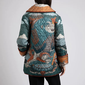 A person wears the SABINA SAVAGE - THE SNOW LION QUILTED JACKET from SABINA SAVAGE ENGLAND, featuring a colorful, intricately designed nature-themed pattern with trees, sky, and animals. Reminiscent of a reversible silk twill jacket, it features a wide collar, contrasting orange inner lining, and large pockets. The person is shown from the shoulders down.